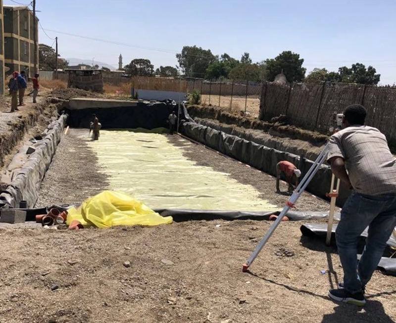 A surveyor setting levels while workers spread gravel and protection cloth in a lined wetland basin